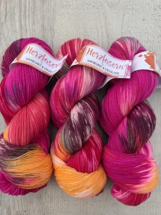 Drei handgefärbte Wollstränge in kräftigem Pink, Orange und dunklen Kontrasten auf hellem Holzuntergrund, Garnbasis Herzfasern No. 20